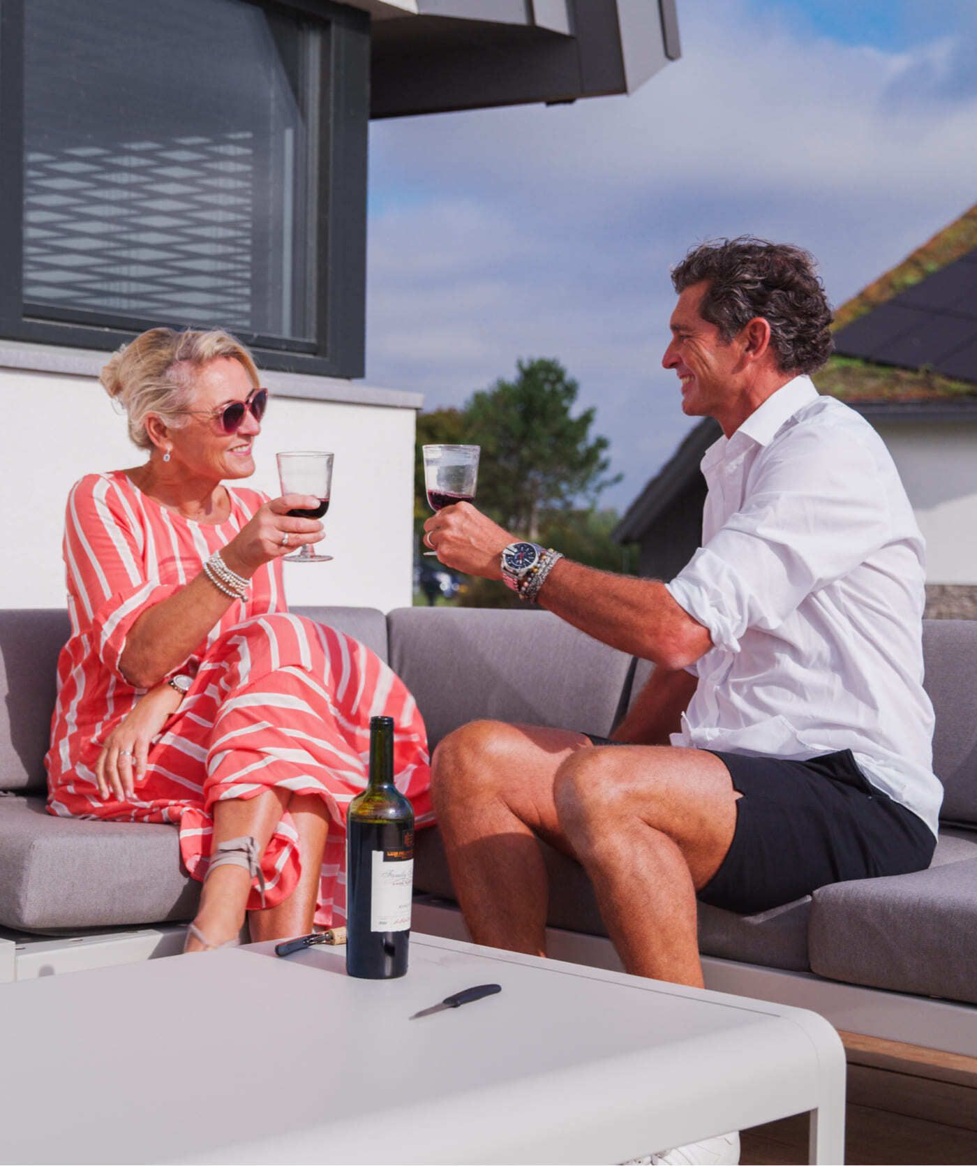 Twee mensen op een buitenbank klinken met hun glazen en glimlachen naar elkaar. Een fles wijn, een telefoon met webshops en een relaxte sfeer bepalen de sfeer op tafel voor hen.