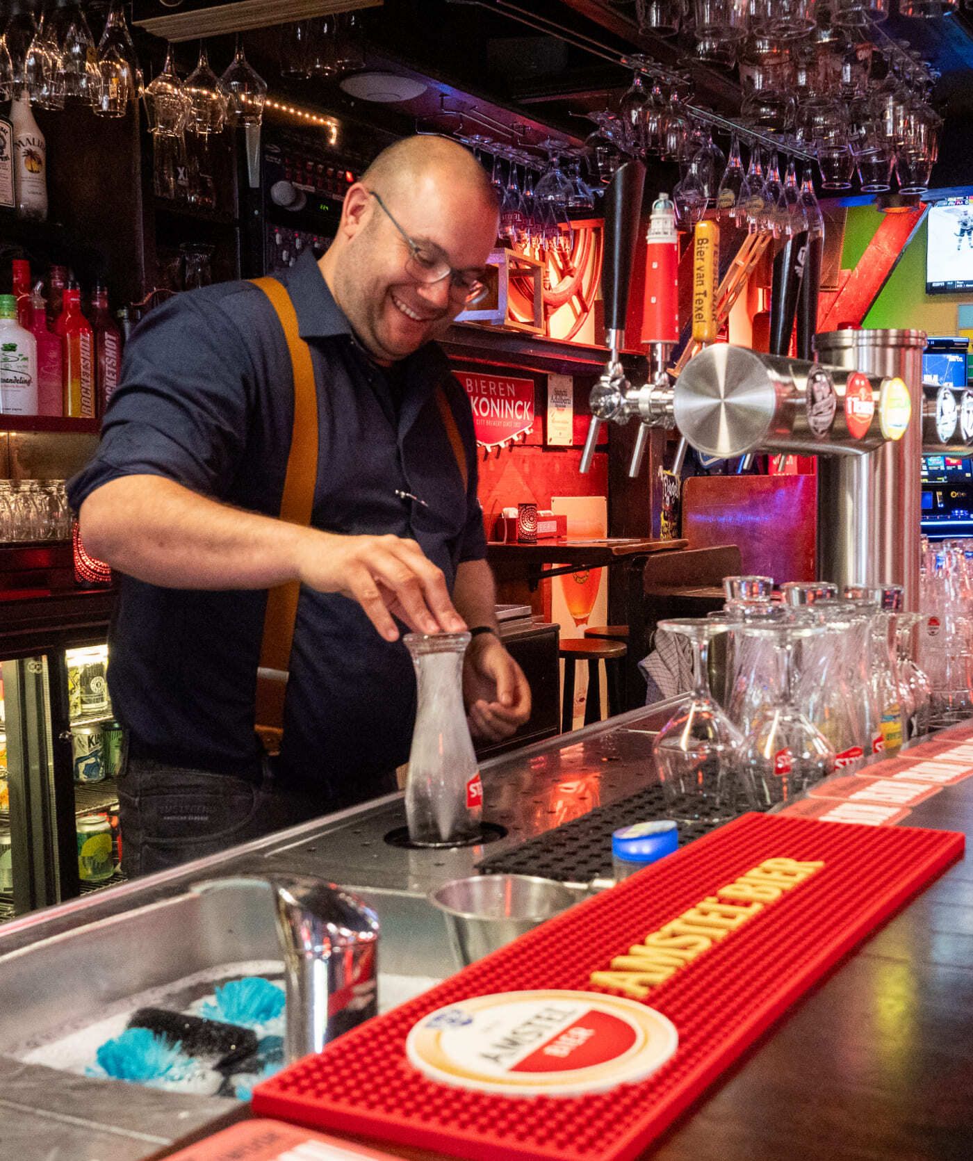 Een barman in een marineblauw overhemd en bretels glimlacht terwijl hij achter een goed gevulde bar een drankje klaarmaakt, terwijl klanten onder het genot van een cocktail praten over websites en online diensten.