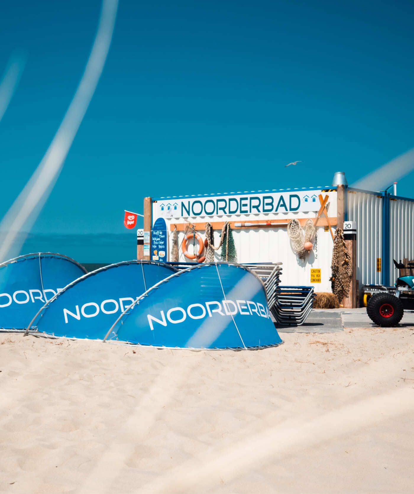 Een strandtafereel met blauwe tenten met het opschrift 'Noorderbad' voor een klein gebouw aan het strand, eveneens met het opschrift 'Noorderbad', onder een helderblauwe lucht. De ideale plek om te ontspannen of om webshops en online diensten te bekijken.