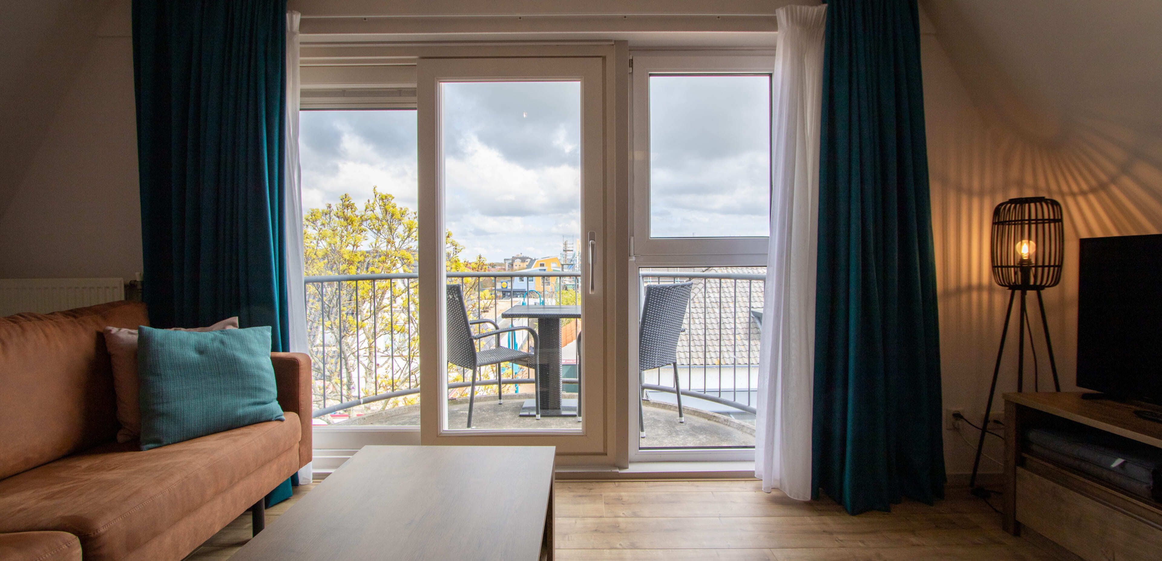 Woonkamer met bruine bank, blauwgroen kussen, salontafel en staande lamp, met uitzicht op een glazen deur naar een klein balkon met twee stoelen en uitzicht op de stad – perfecte inspiratie voor webshops of websites die online diensten aanbieden.