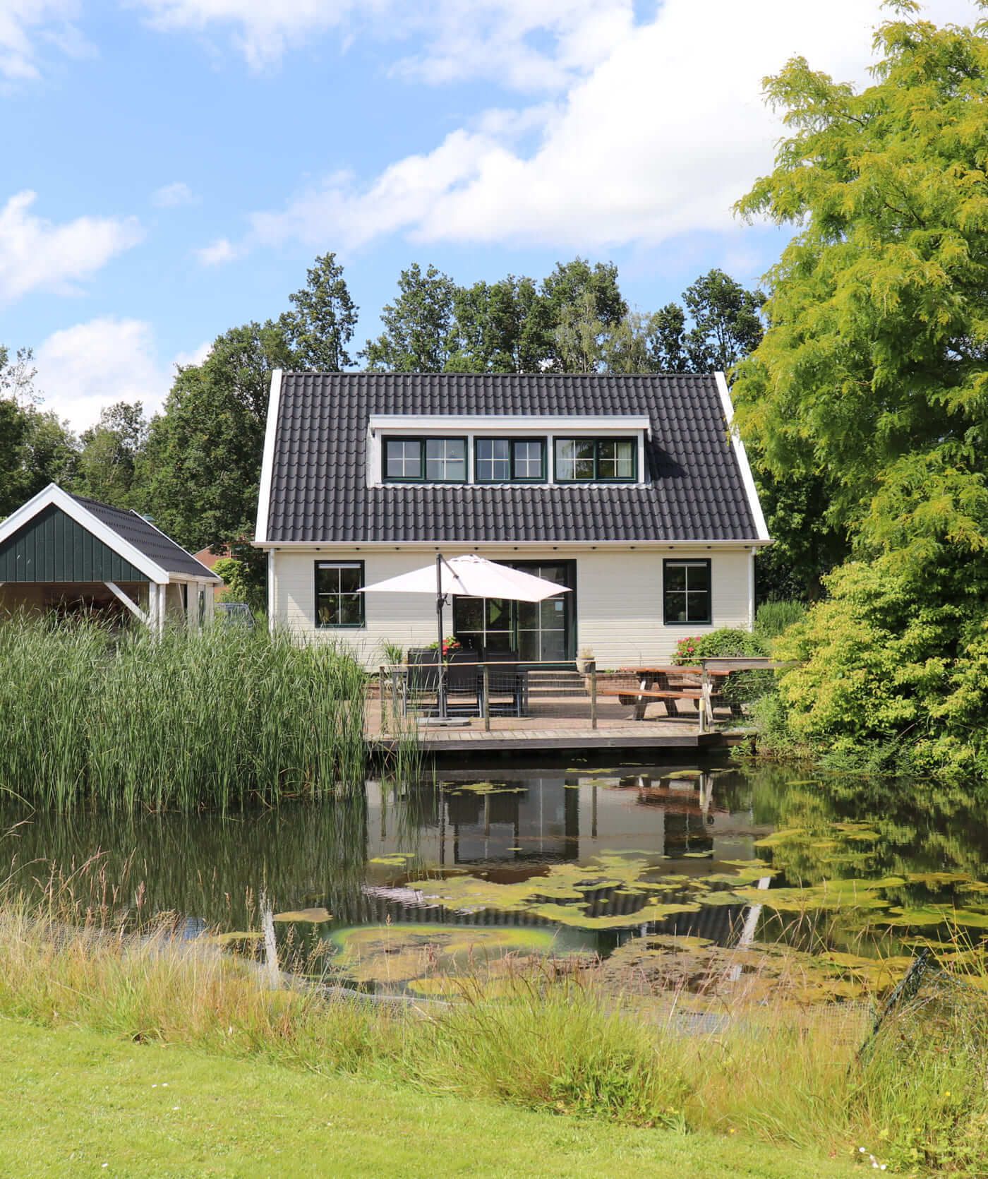 Een wit huis met twee verdiepingen en een donker dak staat naast een kleine vijver, met een houten terras met een tafel en parasol. De perfecte inspiratiebron voor websites die vredige toevluchtsoorden midden in het groen willen tonen.