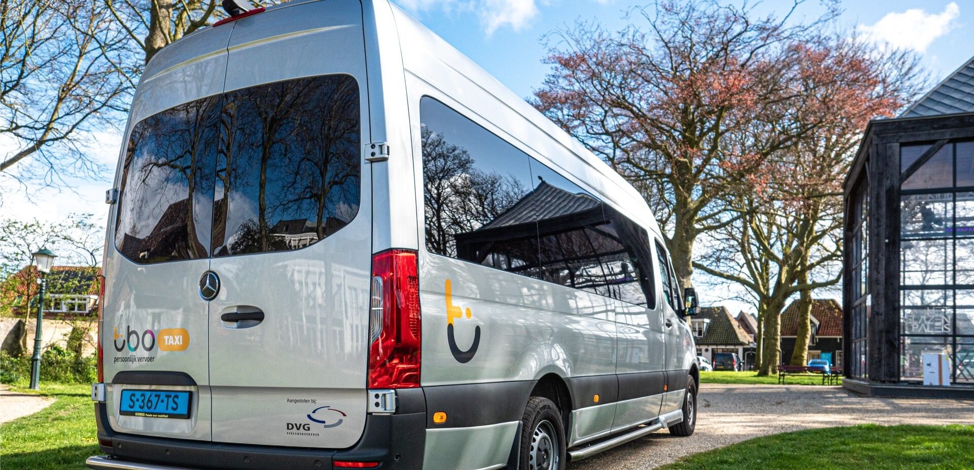Zilveren personenbusje met een blauw kenteken, geparkeerd op een oprit bij een modern glazen gebouw en bomen, ideaal voor bedrijven die online diensten aanbieden of webwinkels die op zoek zijn naar betrouwbaar vervoer.