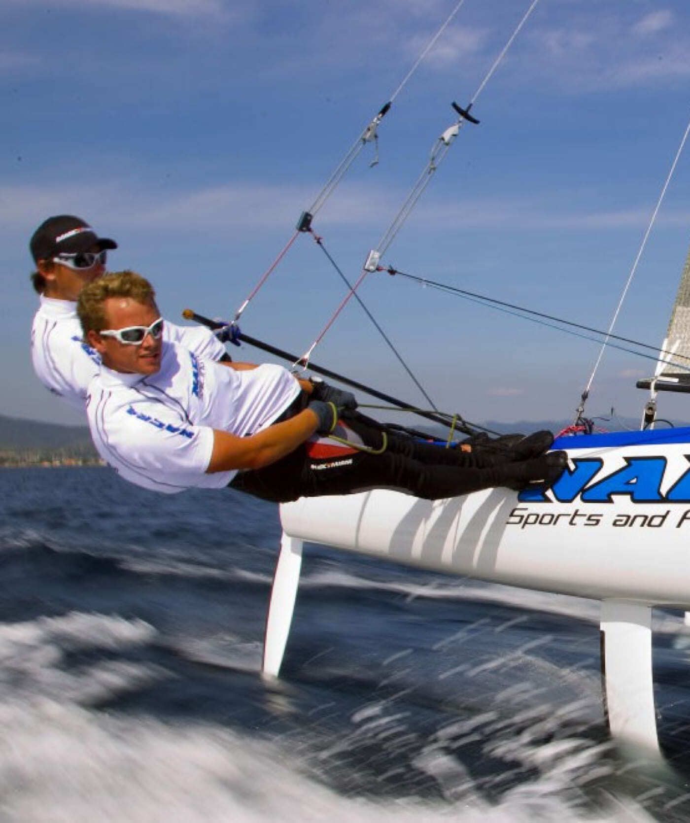 Twee zeilers met zonnebrillen en harnassen leunen over de zijkant van een catamaranzeilboot en bewegen zich snel over het water onder een blauwe lucht - net zoals de beste online diensten ervoor zorgen dat uw webshops of websites soepel blijven draaien.