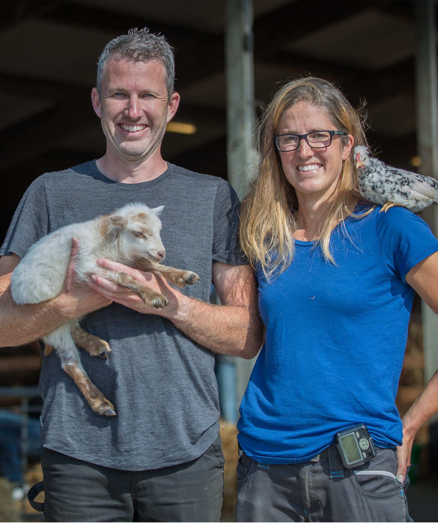 Twee mensen staan naast elkaar bij een schuur; de een houdt een jonge witte geit vast en de ander heeft een vogel op zijn schouder. Ze zouden websites of webshops kunnen bespreken terwijl ze van de buitenlucht genieten.